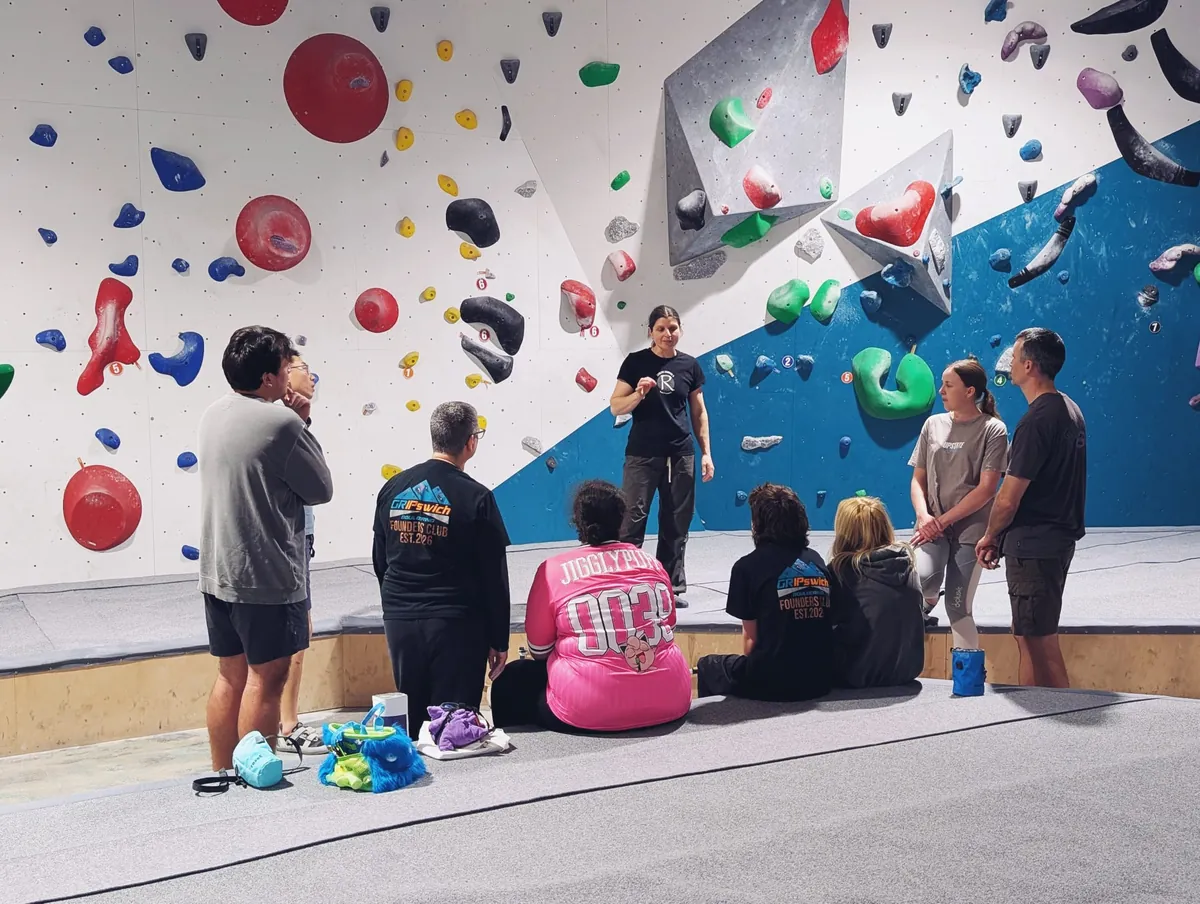 An instructor talking to a Bouldering 101 group at GRIPswich, the class standing and sitting on the mats in front of the blue and white wall